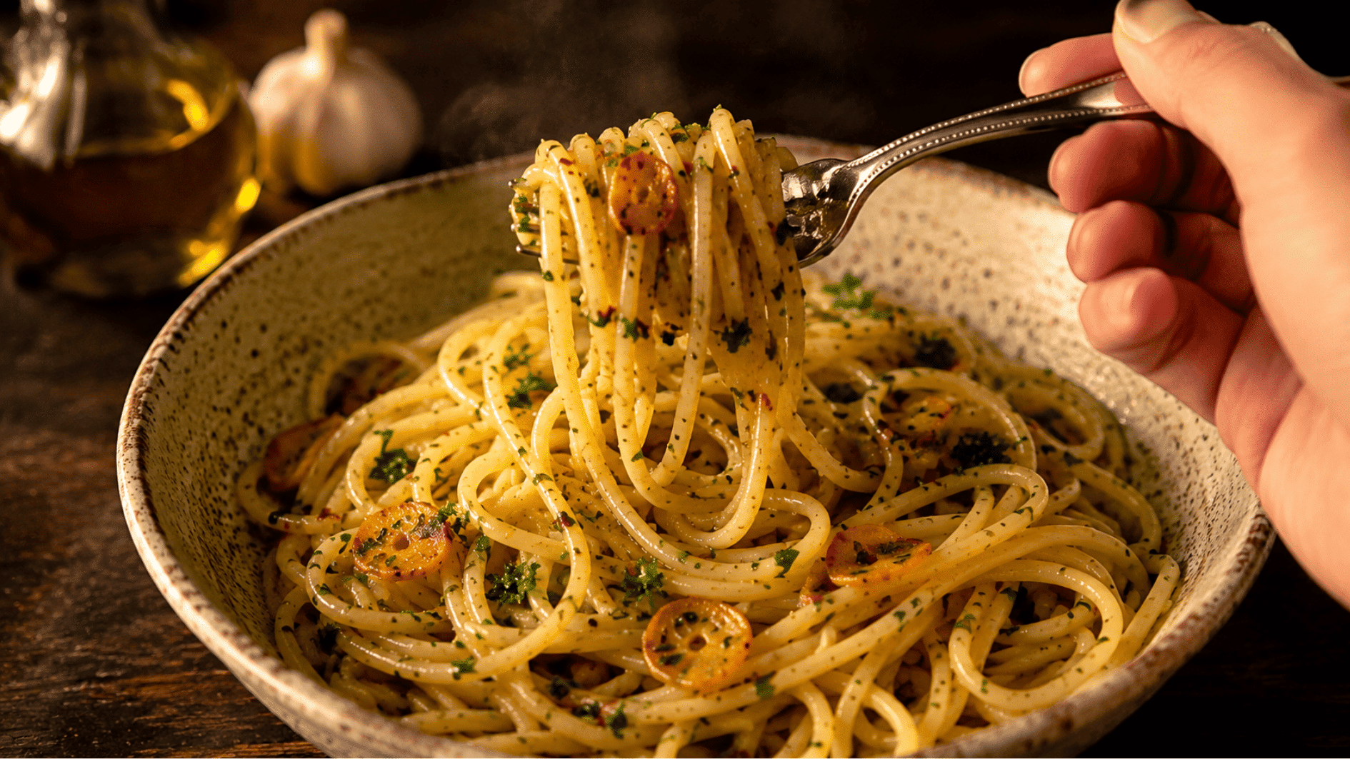 A hand twirling spaghetti on a fork over a rustic bowl with garlic and parsley in warm lighting