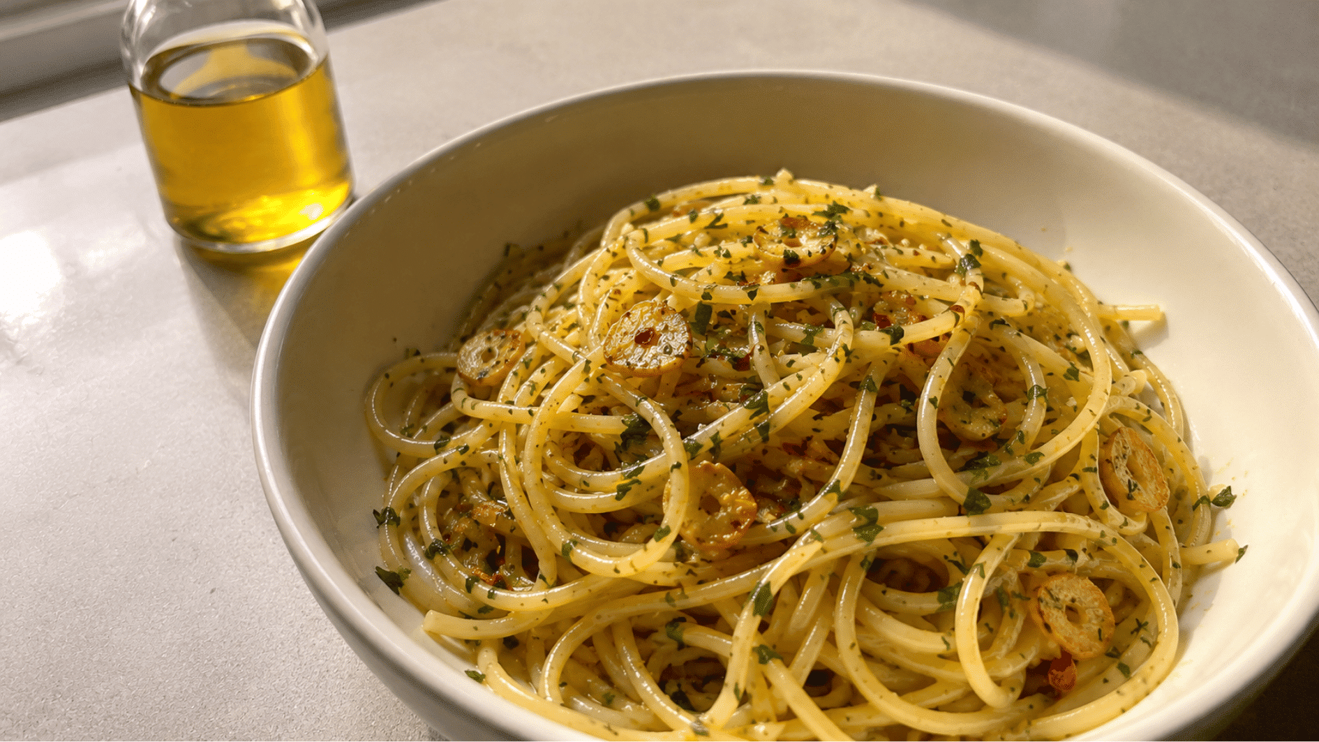 Spaghetti with golden garlic slices, red pepper flakes and parsley beside an olive oil bottle