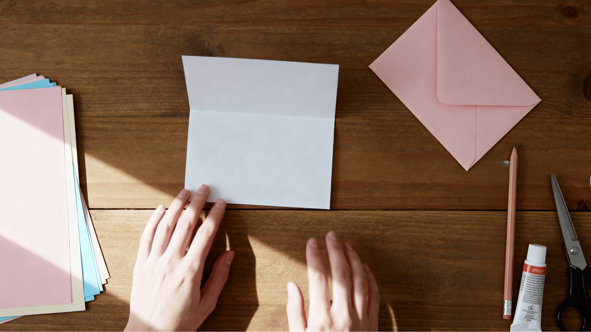 blank white greeting card on wooden craft table with pastel papers, pink envelope, hands, pencil, scissors and glue nearby