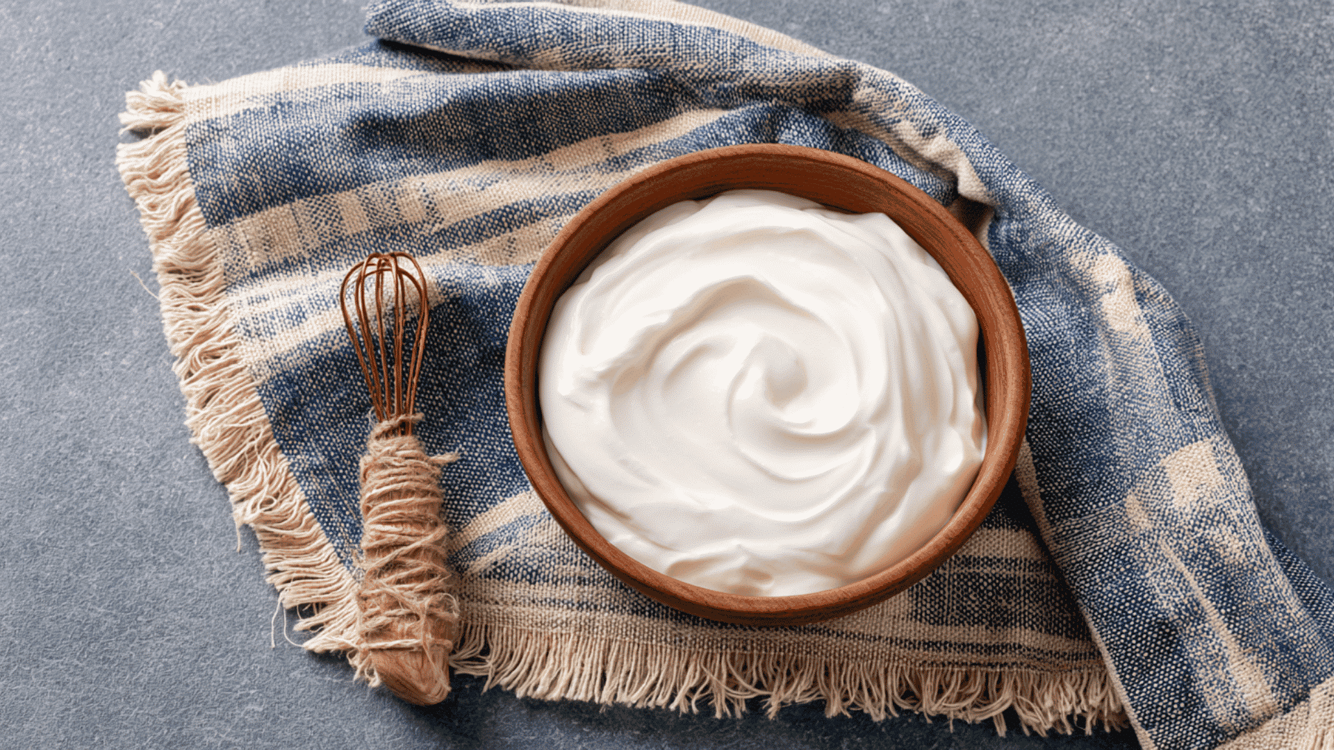 greek style yogurt kept on a grey floor with table cloth behind it with whisker kept on side with visible whisk signs