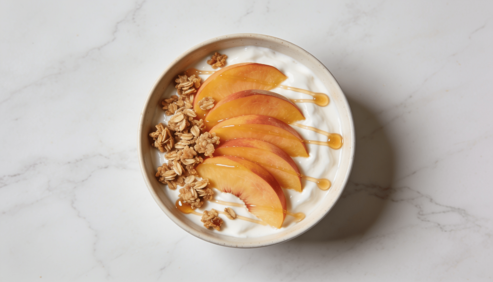 greek yogurt bowl topped with fresh peach slices granola and a light honey drizzle on a marble surface