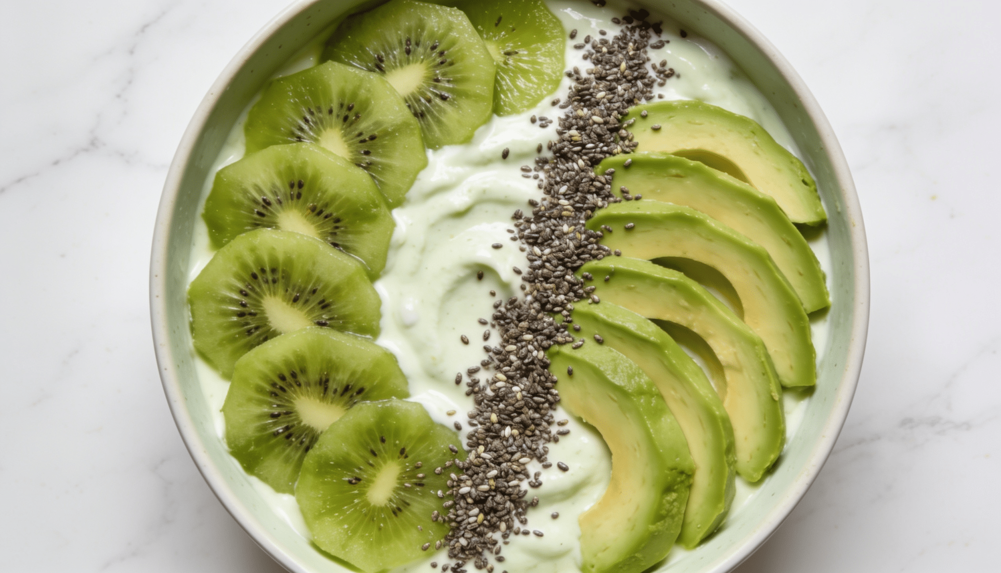 green breakfast bowl with kiwi slices avocado chia seeds and creamy yogurt base in a white bowl on marble surface
