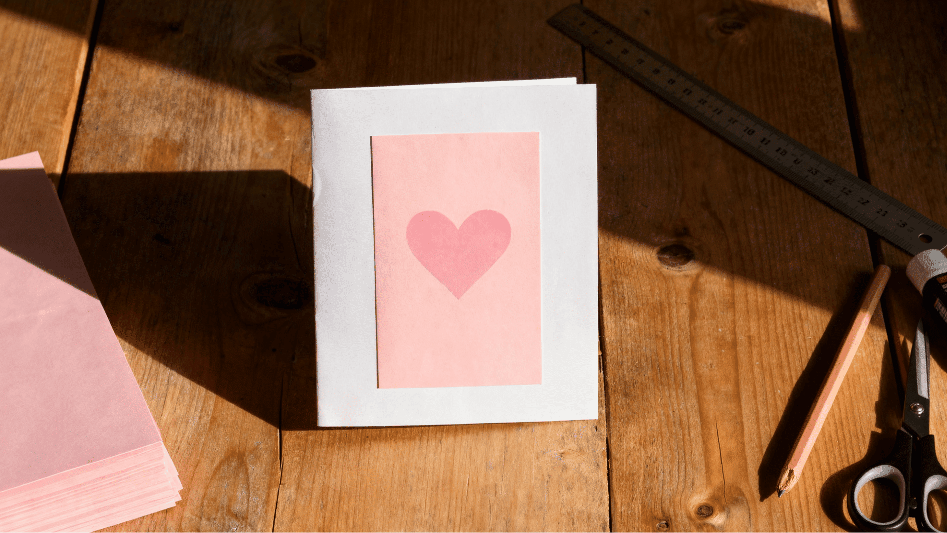 handmade greeting card on wooden craft desk featuring a pink heart on pastel panel, surrounded by scissors, ruler, pencil
