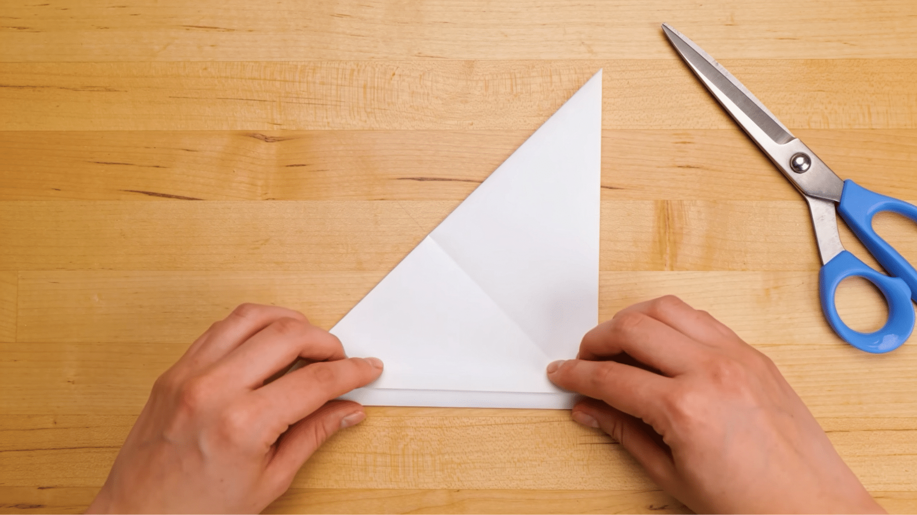 hands folding white paper diagonally to create a square sheet for paper snowflake making