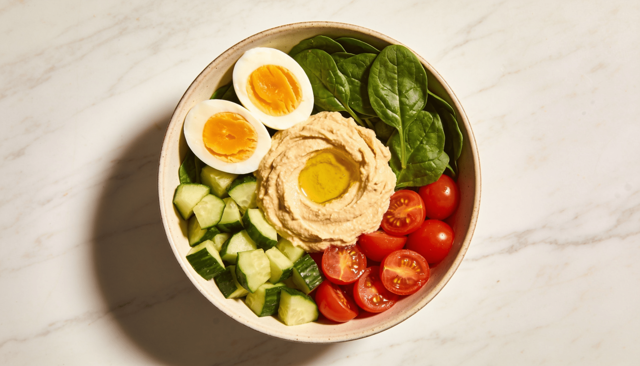 mediterranean breakfast bowl with hummus hard boiled eggs cucumber tomatoes and spinach in a ceramic bowl on marble surface