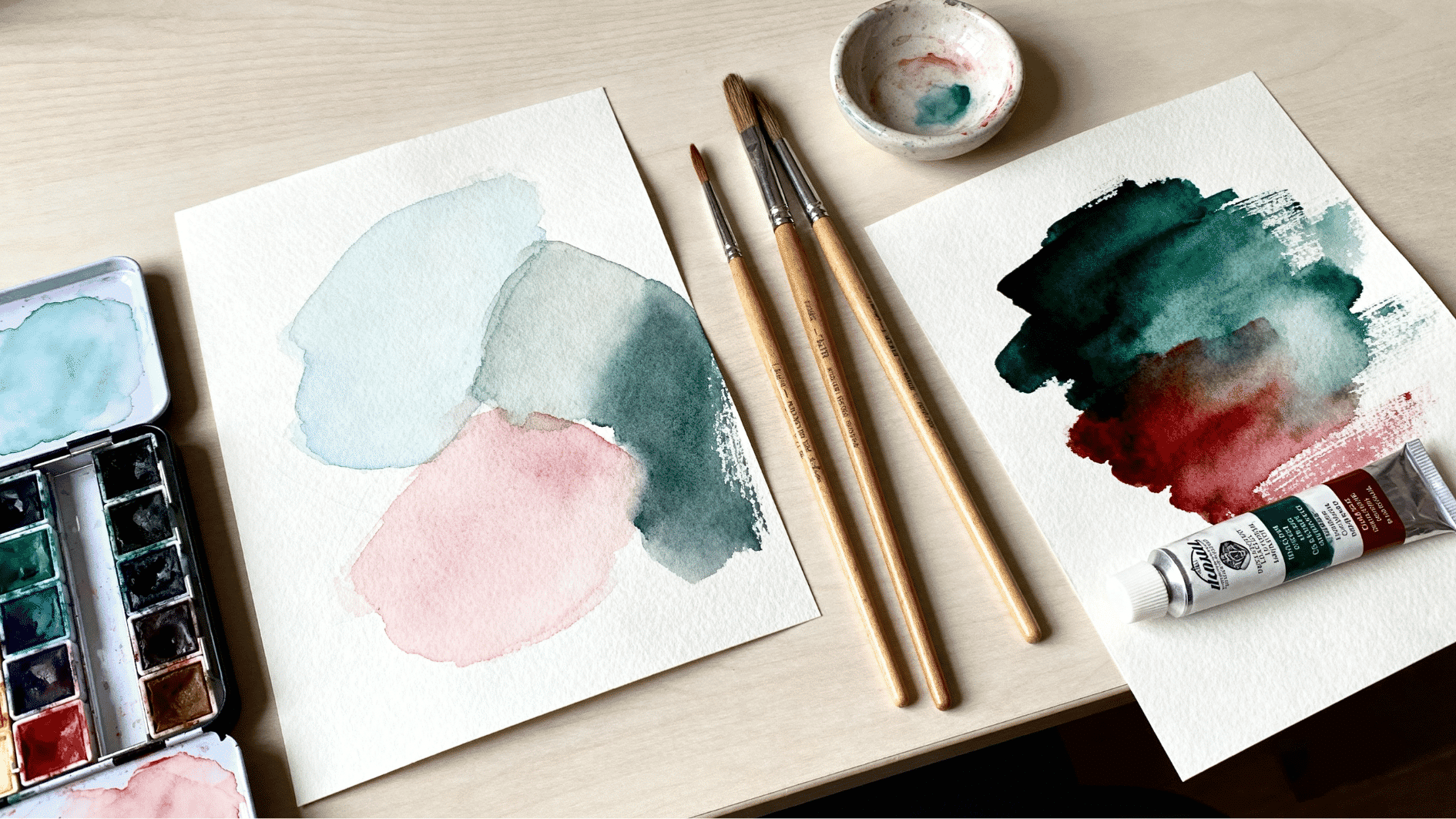 pan and tube watercolor paints shown side by side with brushes, paper, and soft color washes on a desk.