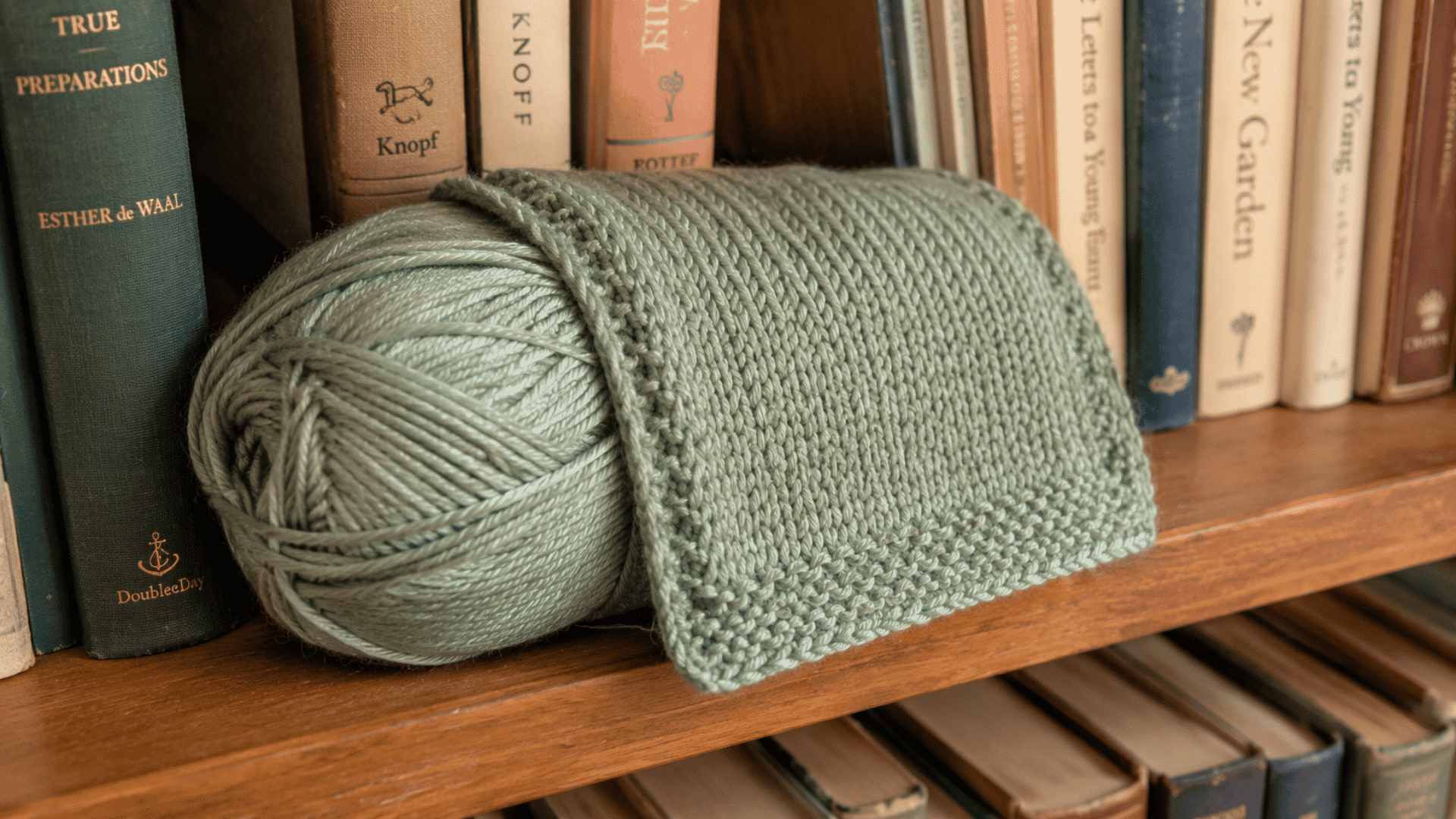 sage green yarn skein with a knitted swatch draped over it sitting on a wooden bookshelf between books