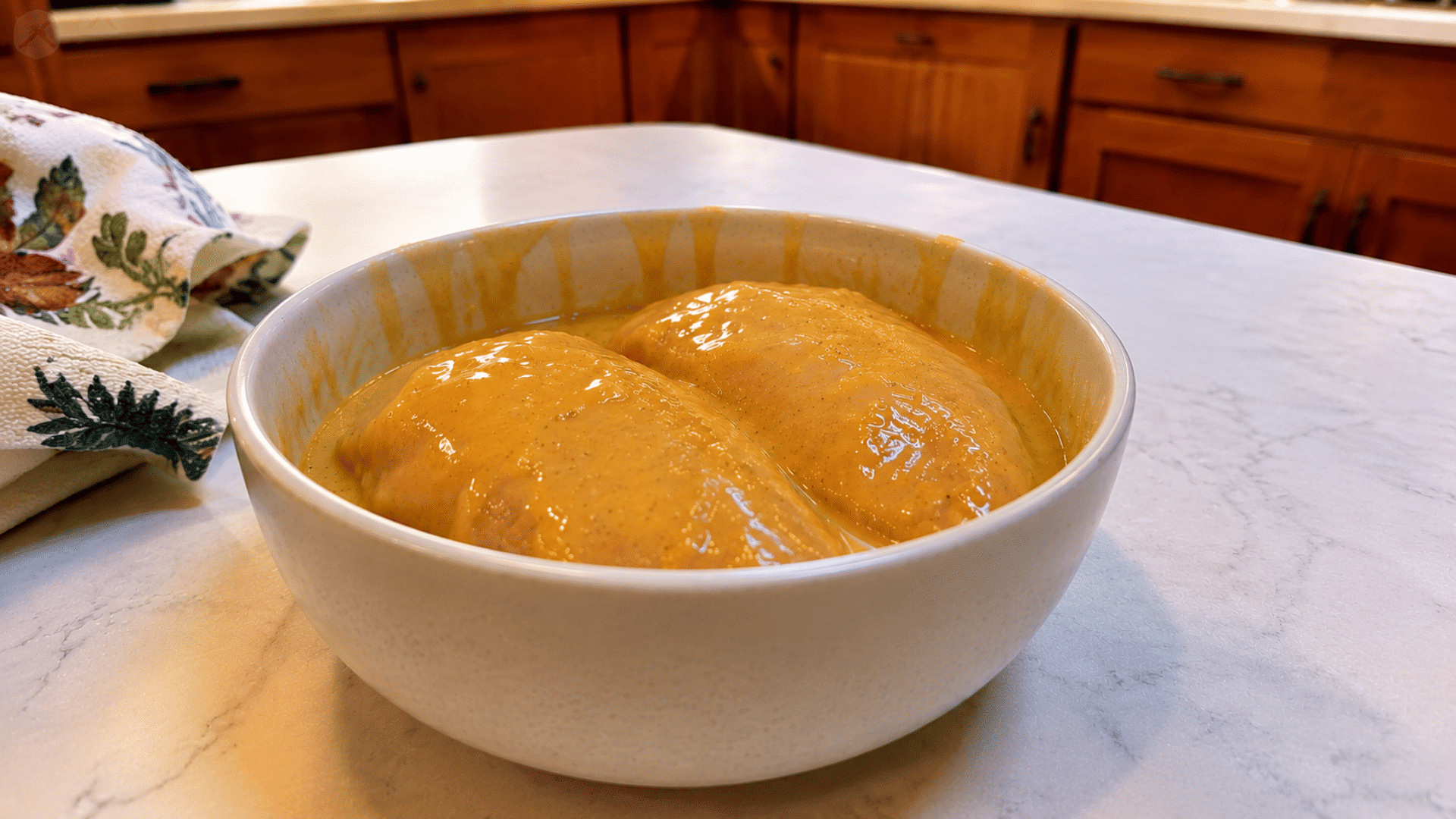 two raw chicken breasts coated in golden honey mustard sauce in a white ceramic bowl