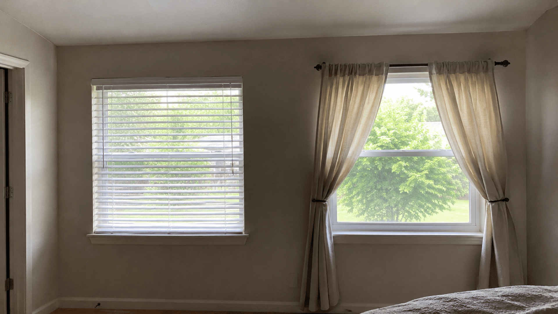 a bedroom with two windows, one fitted with white horizontal blinds and one with sheer linen curtains tied back, natural light coming through both.