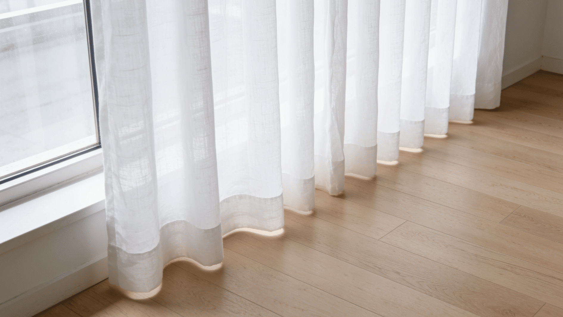 a bright, modern living room with sheer white linen curtains hanging perfectly straight, hem just grazing a light hardwood floor with zero gap and no pooling, soft natural daylight streaming throu