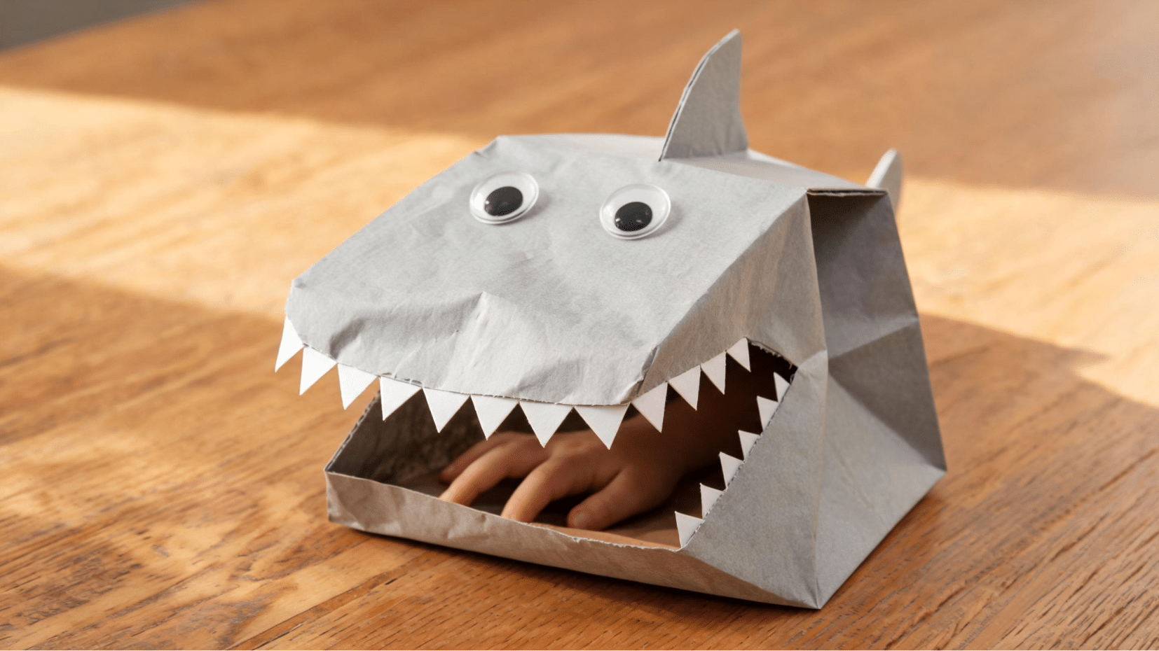 a handmade paper shark puppet with googly eyes on a wooden surface (1)