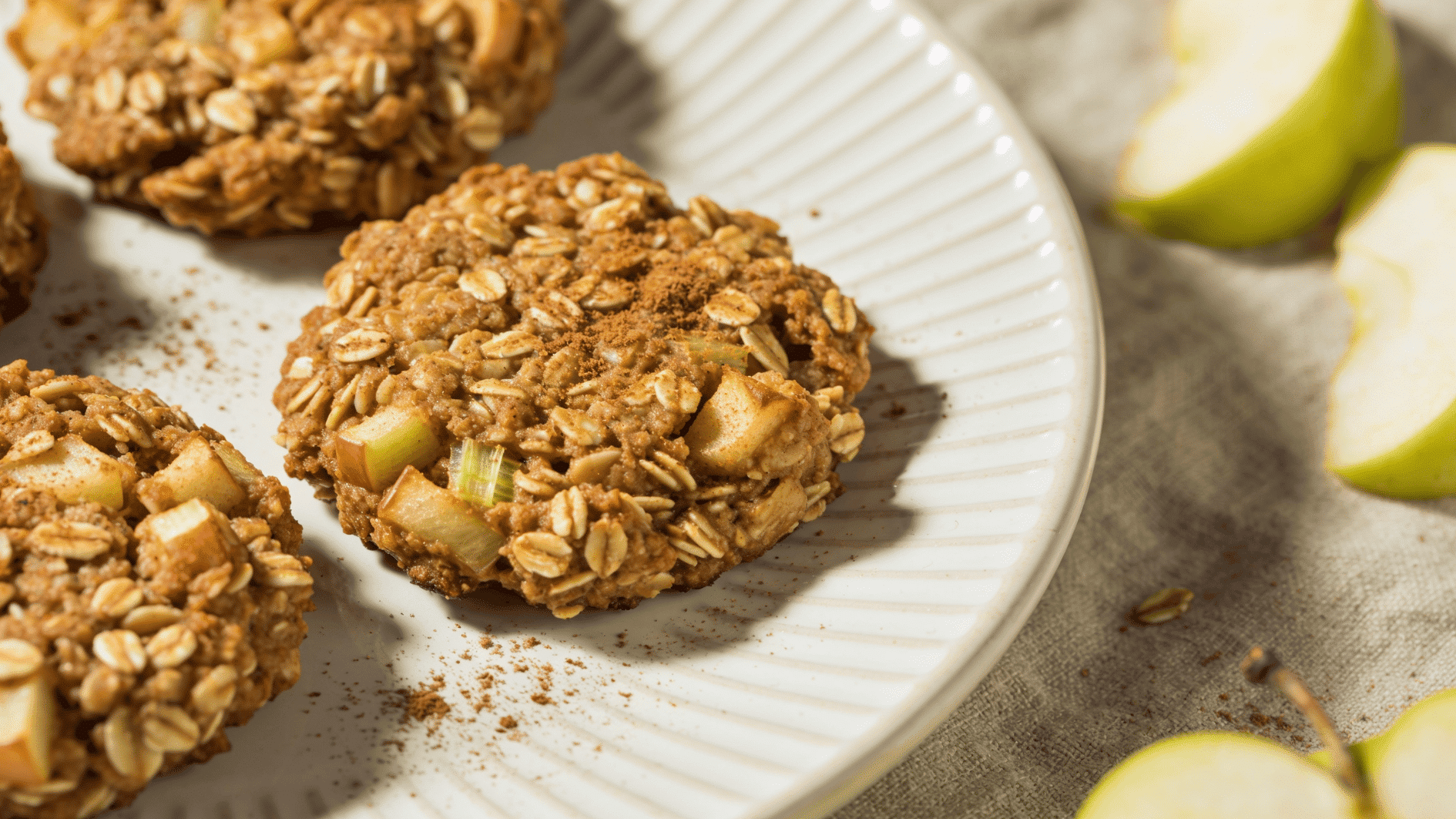 apple cinnamon oat cookies on a white ceramic plate, visible grated apple bits and oats in texture, light golden-brown color, sprinkle of cinnamon dust, small fresh apple slices in background