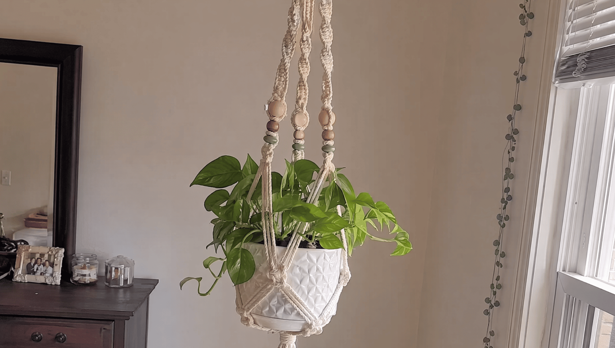 beaded macrame plant hanger with green leaves in a white pot near a sunny home window