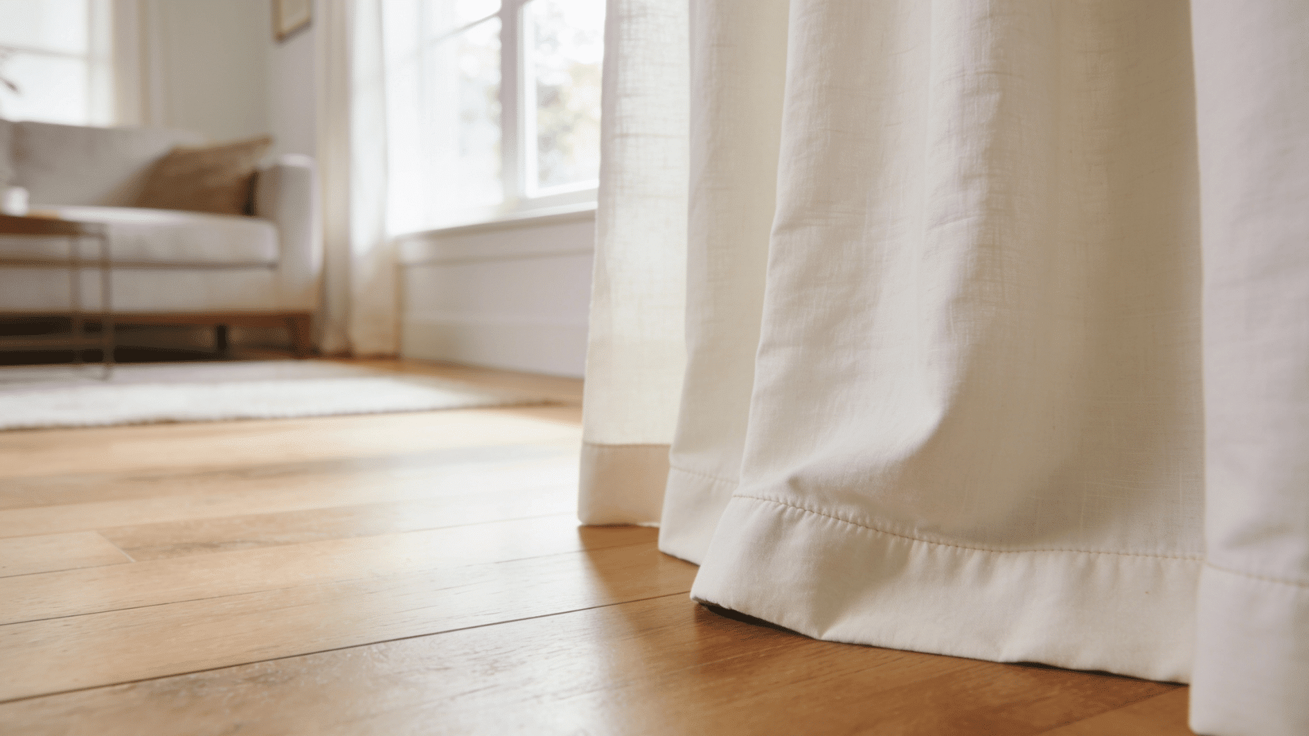 bright, airy living room window with floor-length ivory linen curtains hemmed in a break style — the fabric 1–2 inches too long so the hem folds softly where it meets the wooden floor