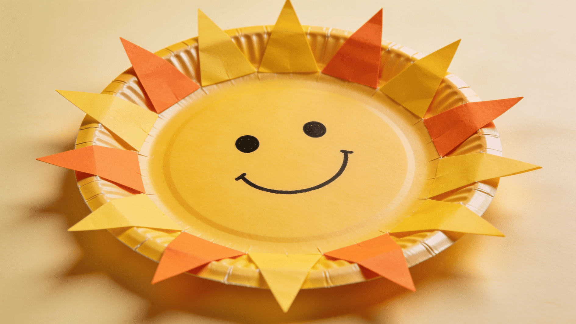 bright yellow and orange paper plate sun craft with triangular paper flares glued around the edges and a simple smiling face in the center