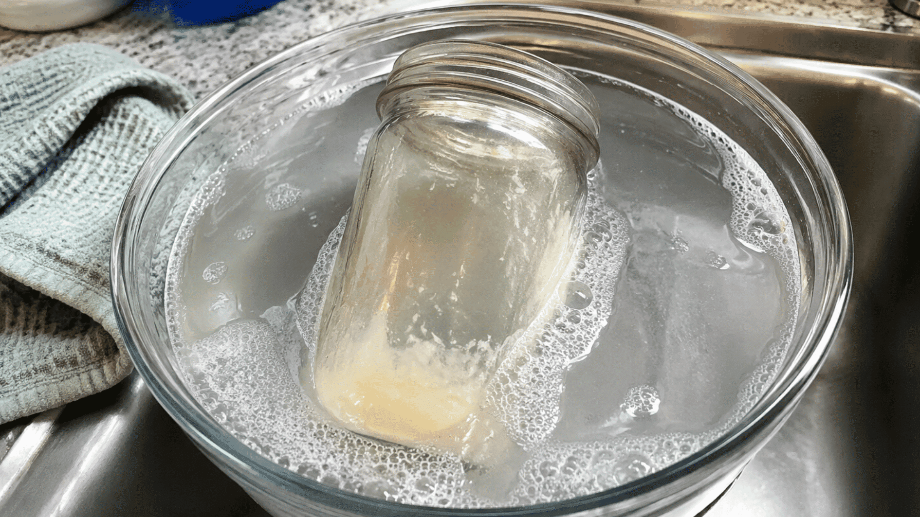 candle jar soaking in warm soapy water with softened wax residue loosening from the glass surface