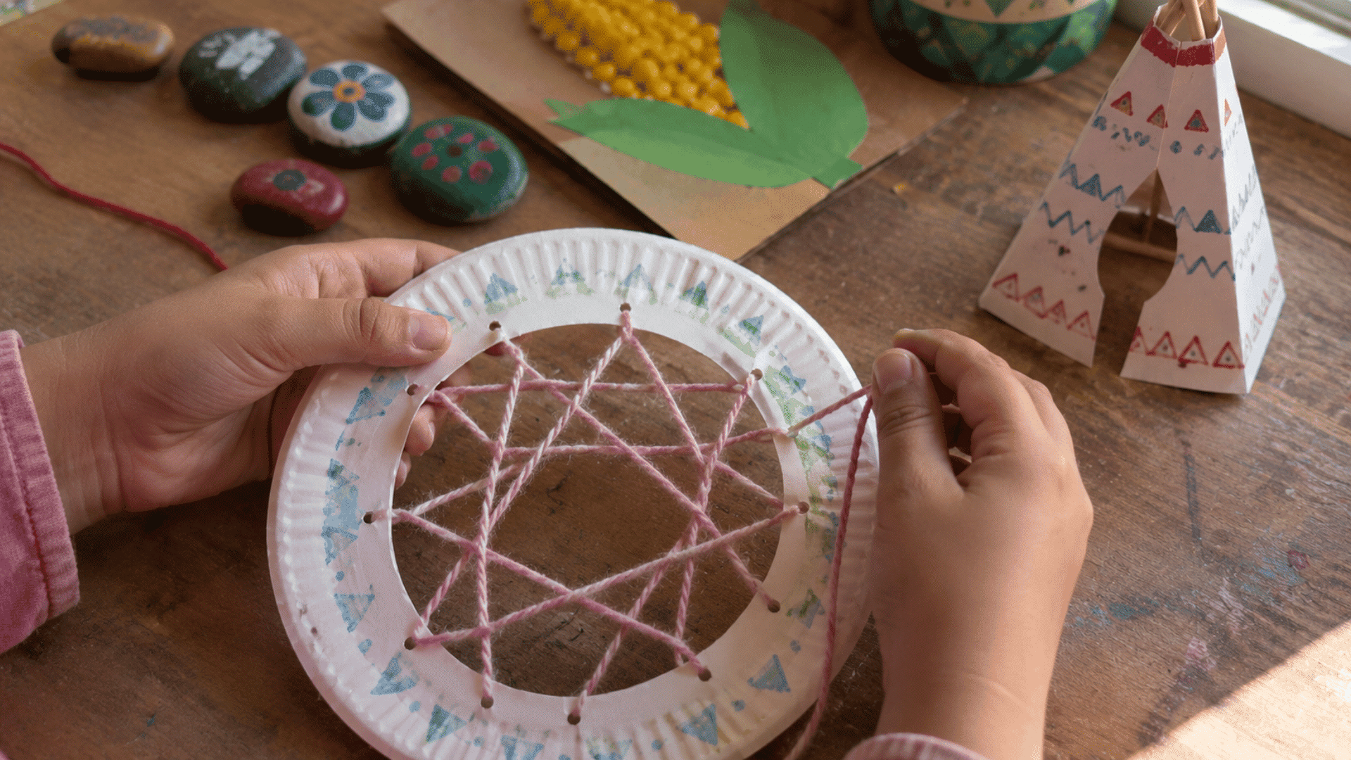 child's hands weaving a paper dreamcatcher surrounded by other native american craft projects