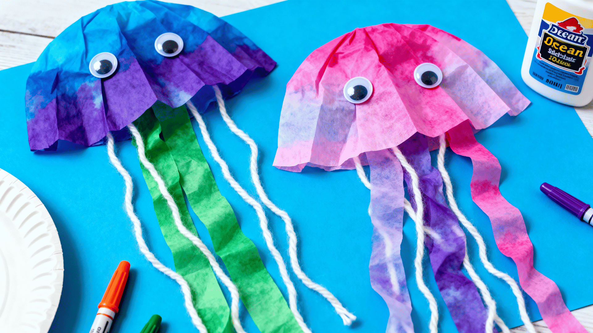 coffee filter jellyfish with yarn tentacles on blue craft paper beside markers, glue, paper plates, and towels