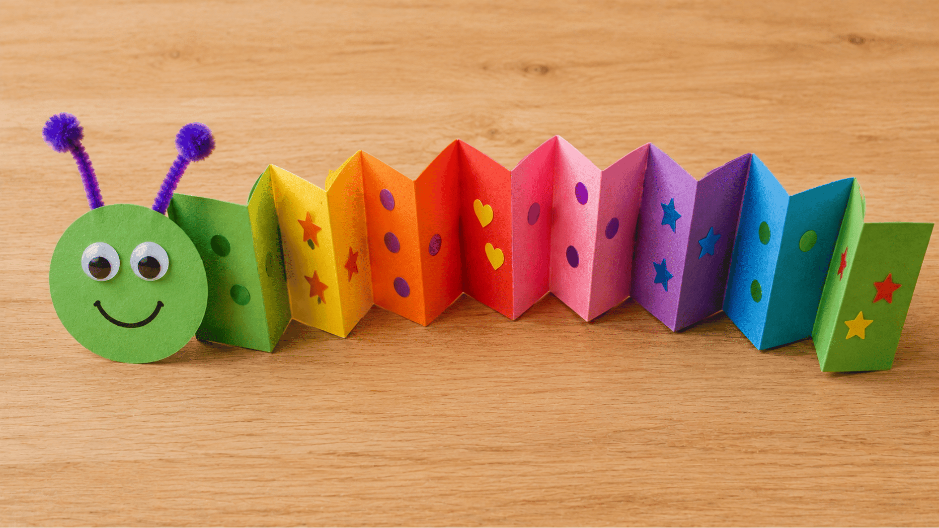 colorful accordion paper caterpillar with a smiling green face and stickers displayed on a wooden table