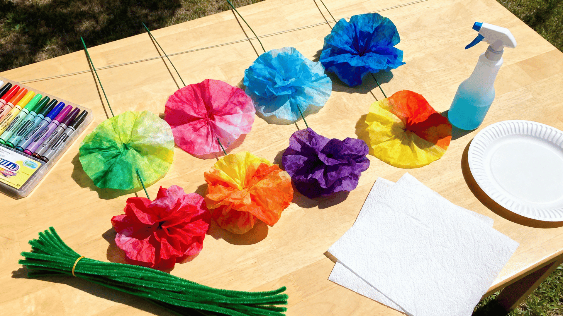 colorful coffee filter flowers with pipe cleaner stems drying on a craft table with markers and paper towels nearby