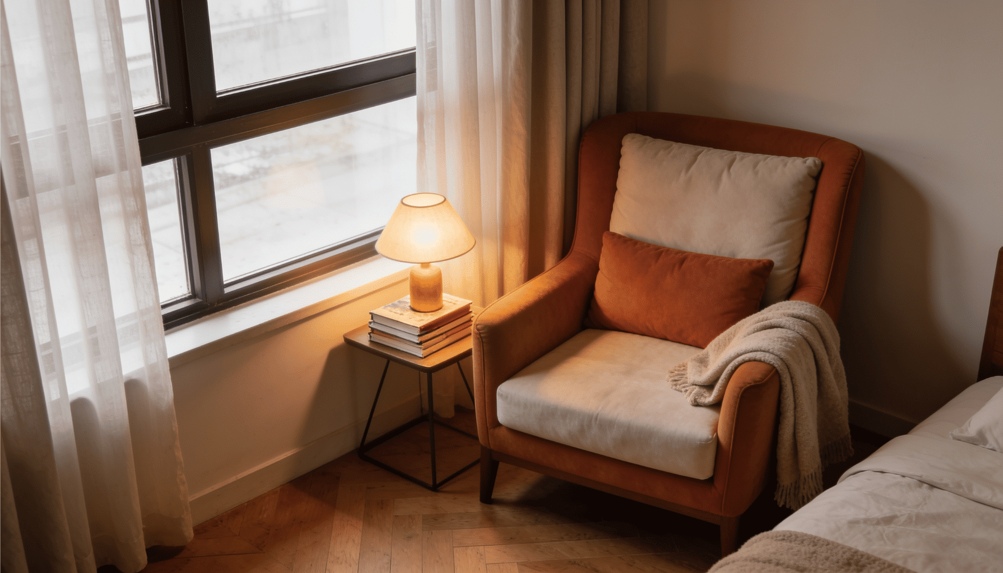 cozy bedroom reading nook with orange armchair, throw blanket, lit lamp, and books beside a bright window