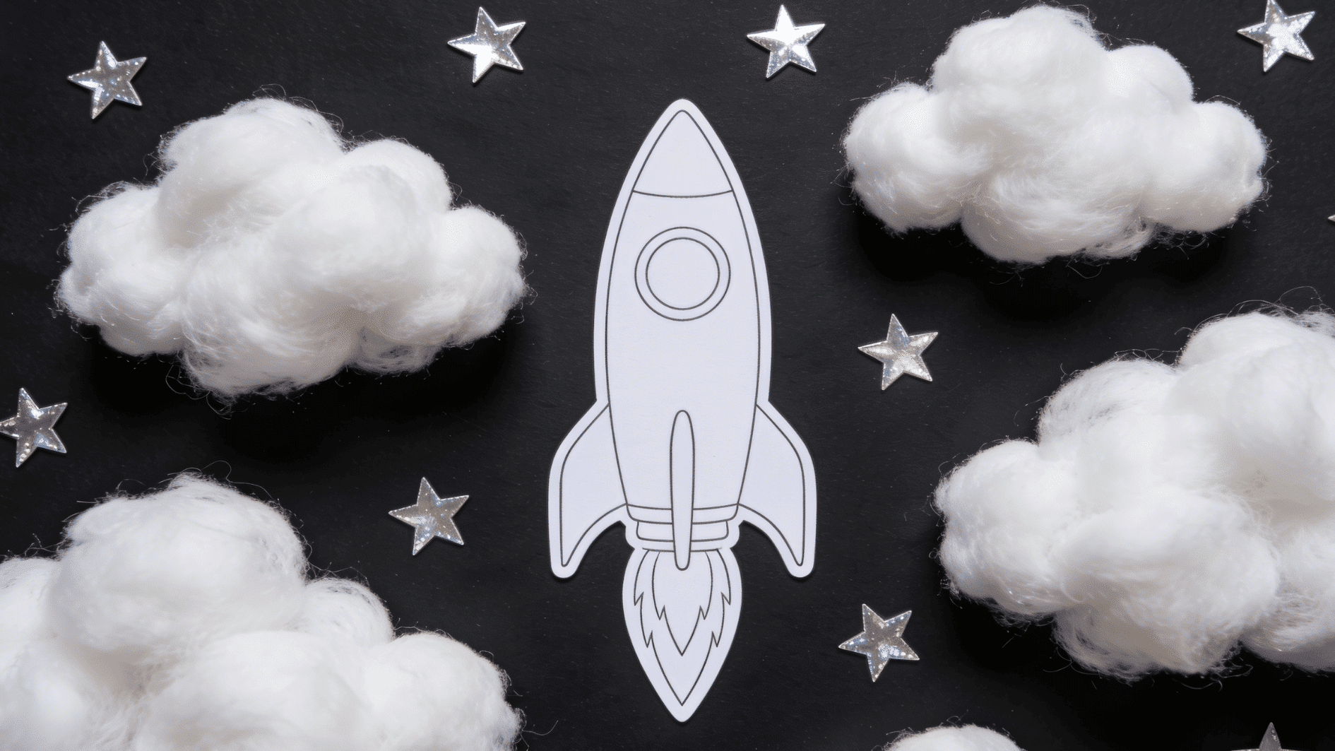 craft scene on black cardstock featuring a printed rocket template surrounded by fluffy cotton ball clouds and star stickers scattered across the dark background