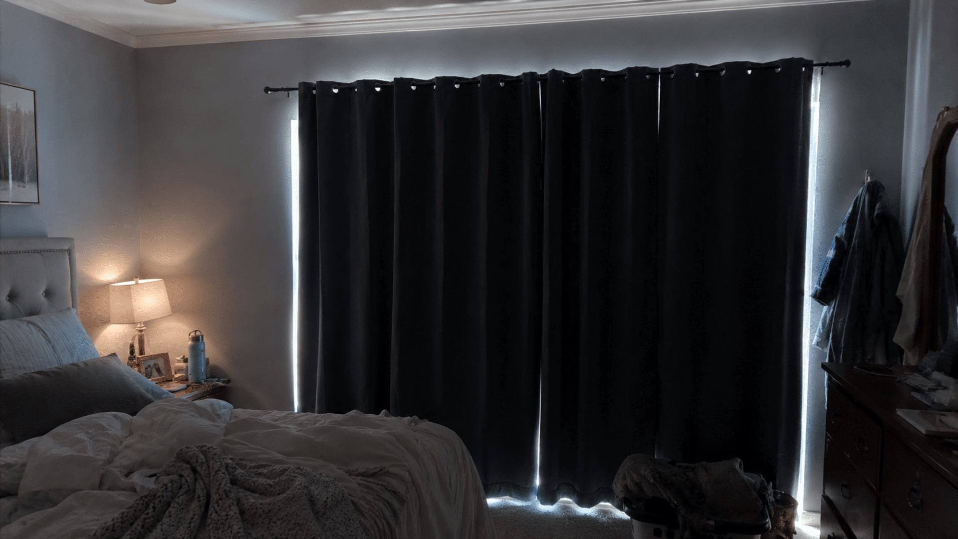 dark bedroom with thick black blackout curtains fully closed, bright daylight leaking around the edges and warm lamp glow