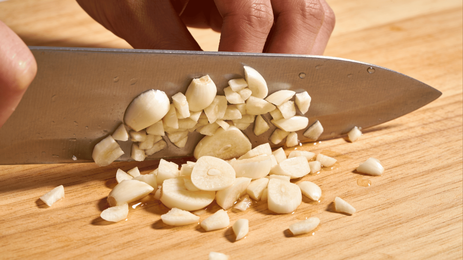 finely minced garlic on a wooden cutting board beside peeled cloves and a sharp kitchen knife