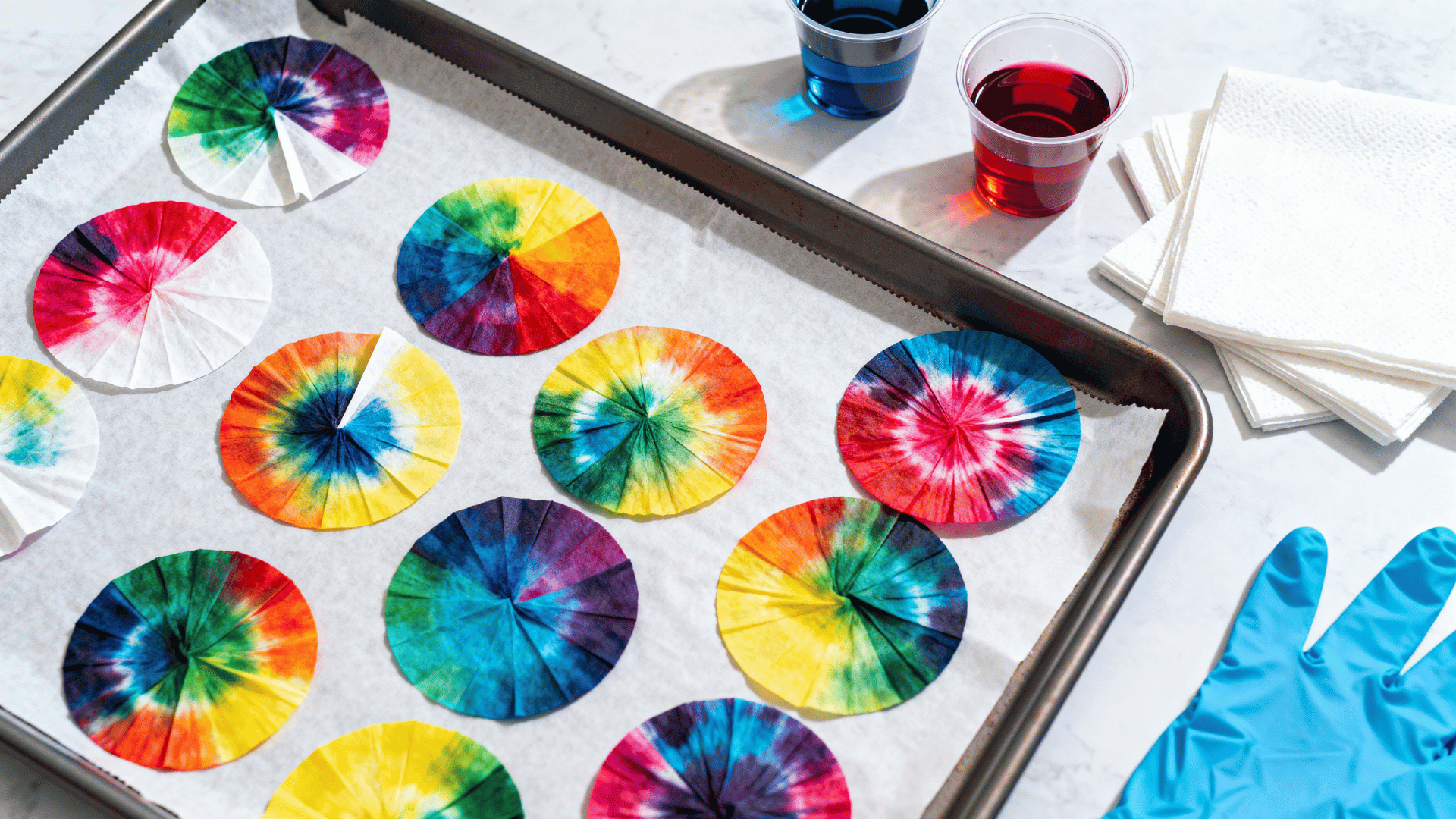 folded coffee filter tie-dye circles opened on a tray beside small cups of liquid color and paper towels nearby