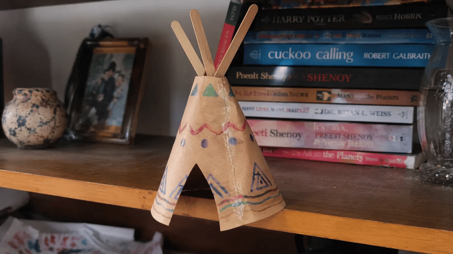 handmade brown paper teepee craft with marker patterns and wooden sticks on a bookshelf