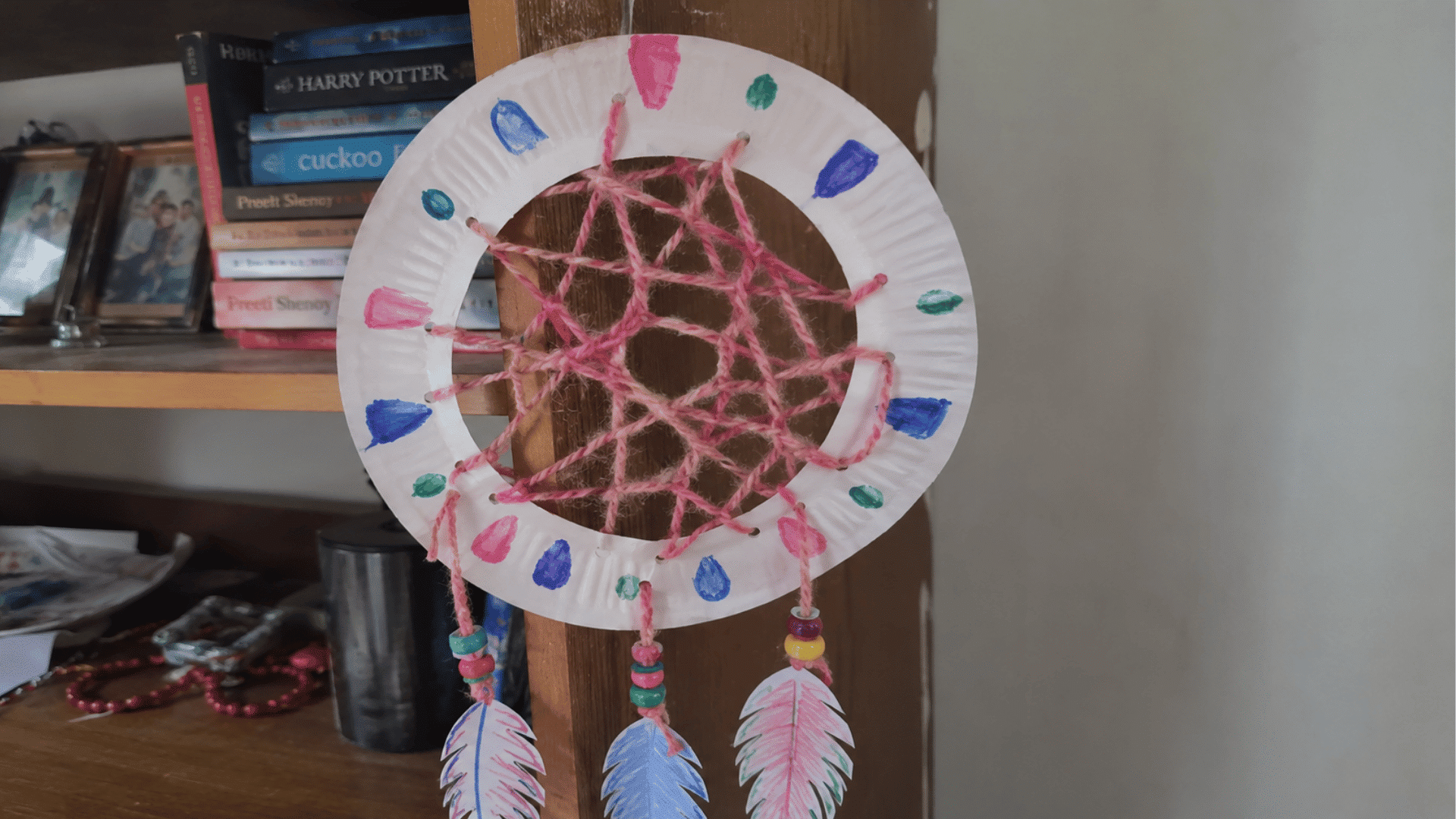 handmade paper plate dreamcatcher with pink yarn web, painted feathers and beads on bookshelf