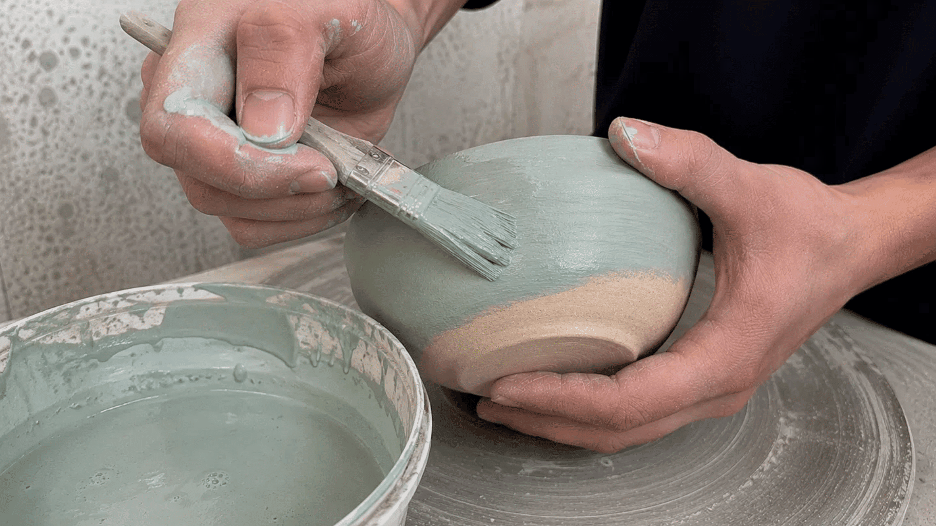 Hands brushing pale green liquid glaze onto a bisque-fired clay bowl, with a bucket of glaze nearby.