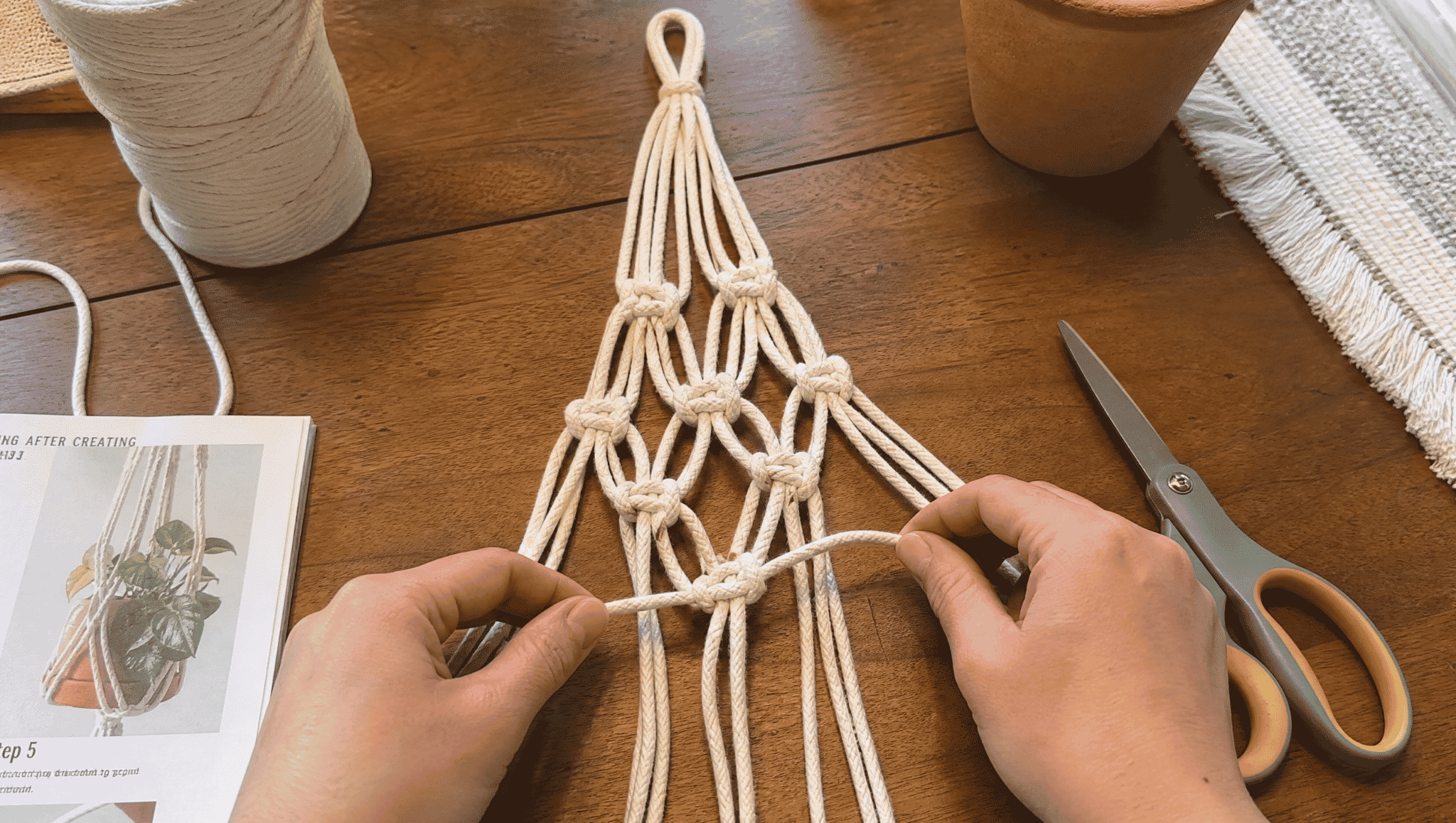 hands creating macrame knots on cords with scissors and materials placed on a wooden table