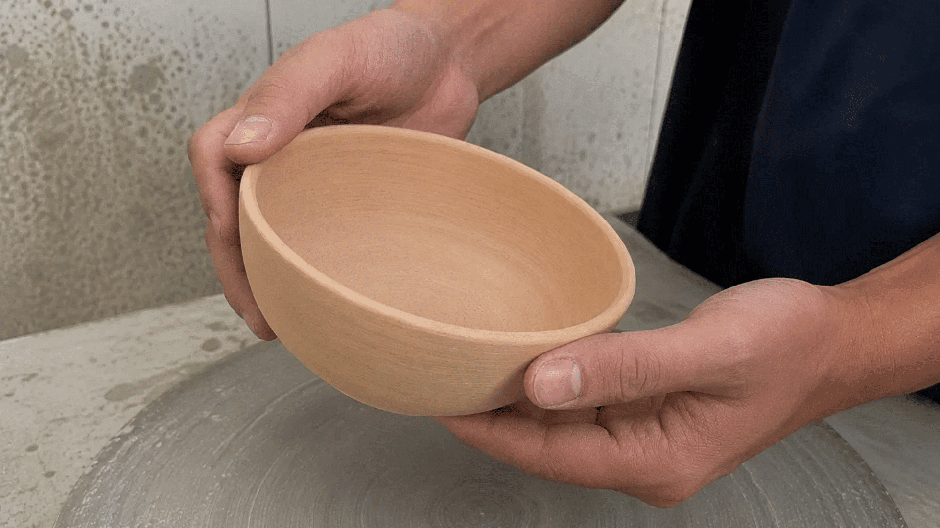 Hands holding up a tilted warm beige bisque-fired clay bowl to show its porous matte surface after firing.