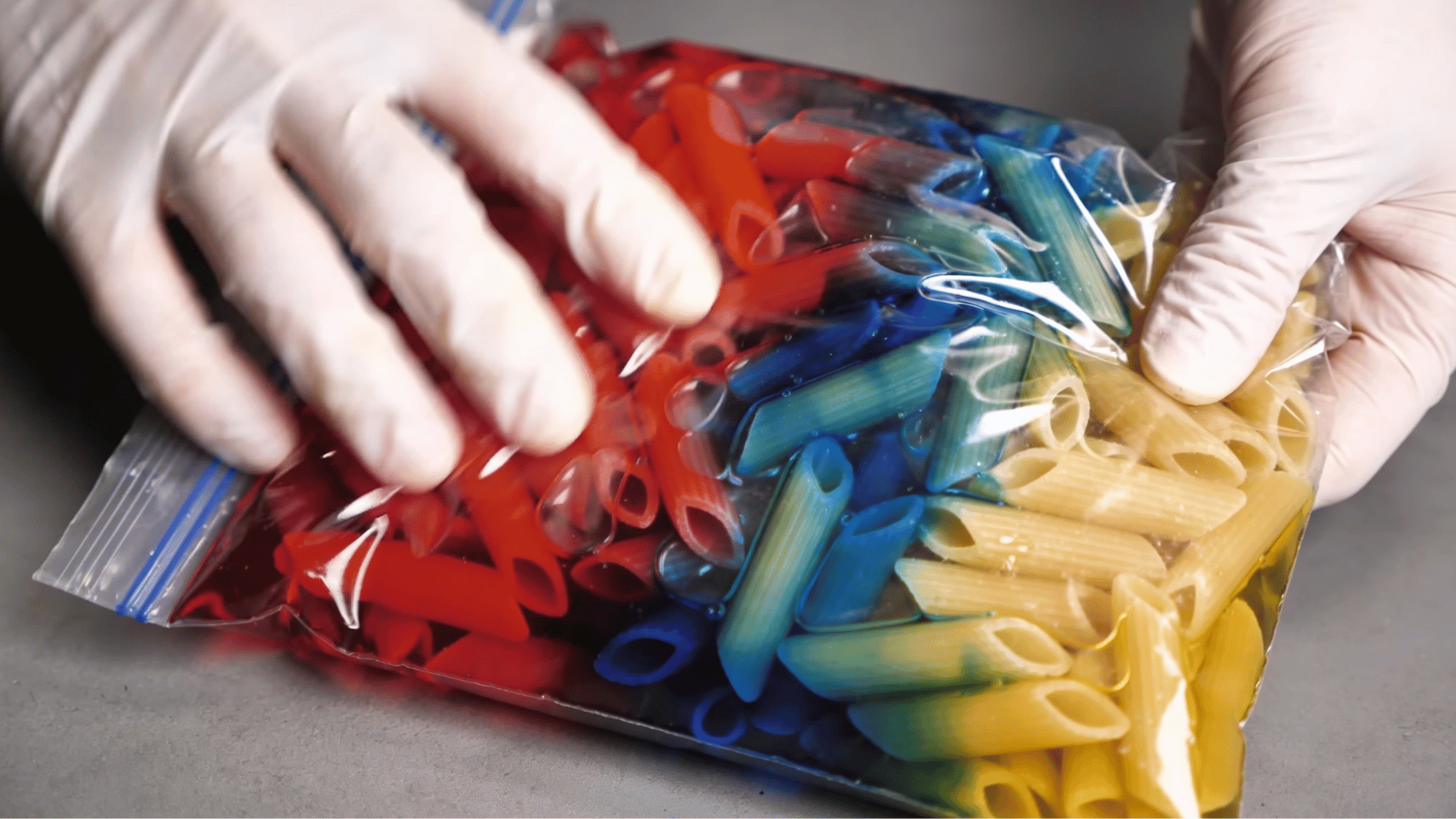 _hands in gloves shaking a zip lock bag with uncooked pasta and bright colored dye on a white countertop