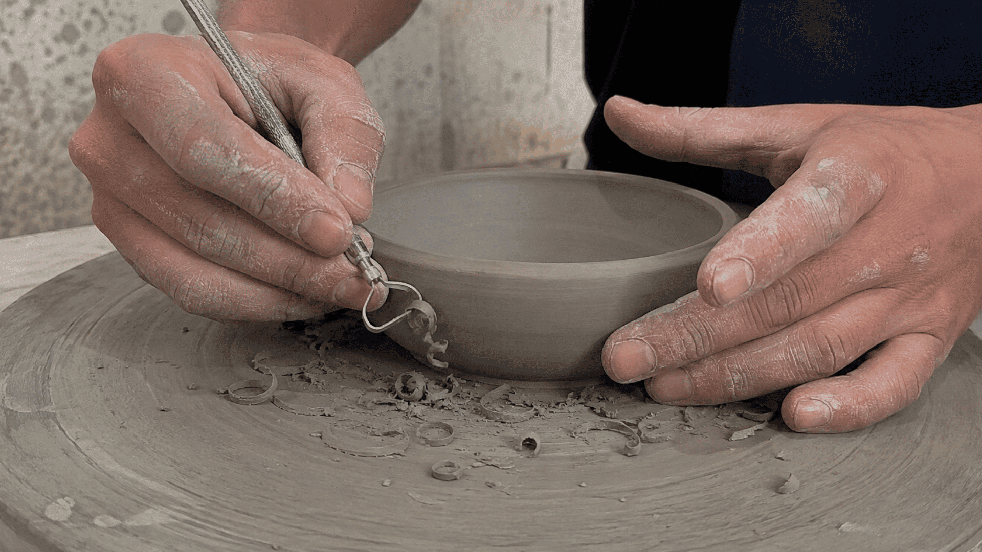 Hands using a trimming tool to carve a firm leather-hard clay bowl, with clay shavings on the pottery wheel.