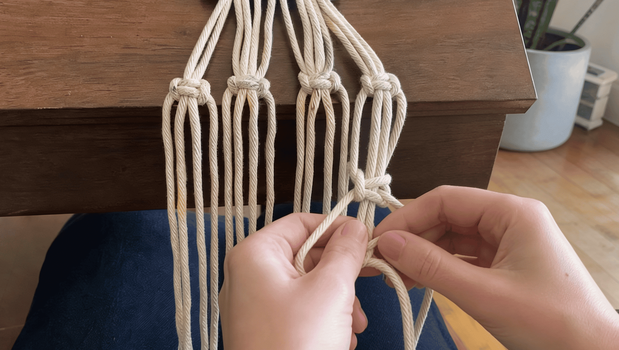 hands weaving macrame cords into a hanging plant holder using a wooden table setup