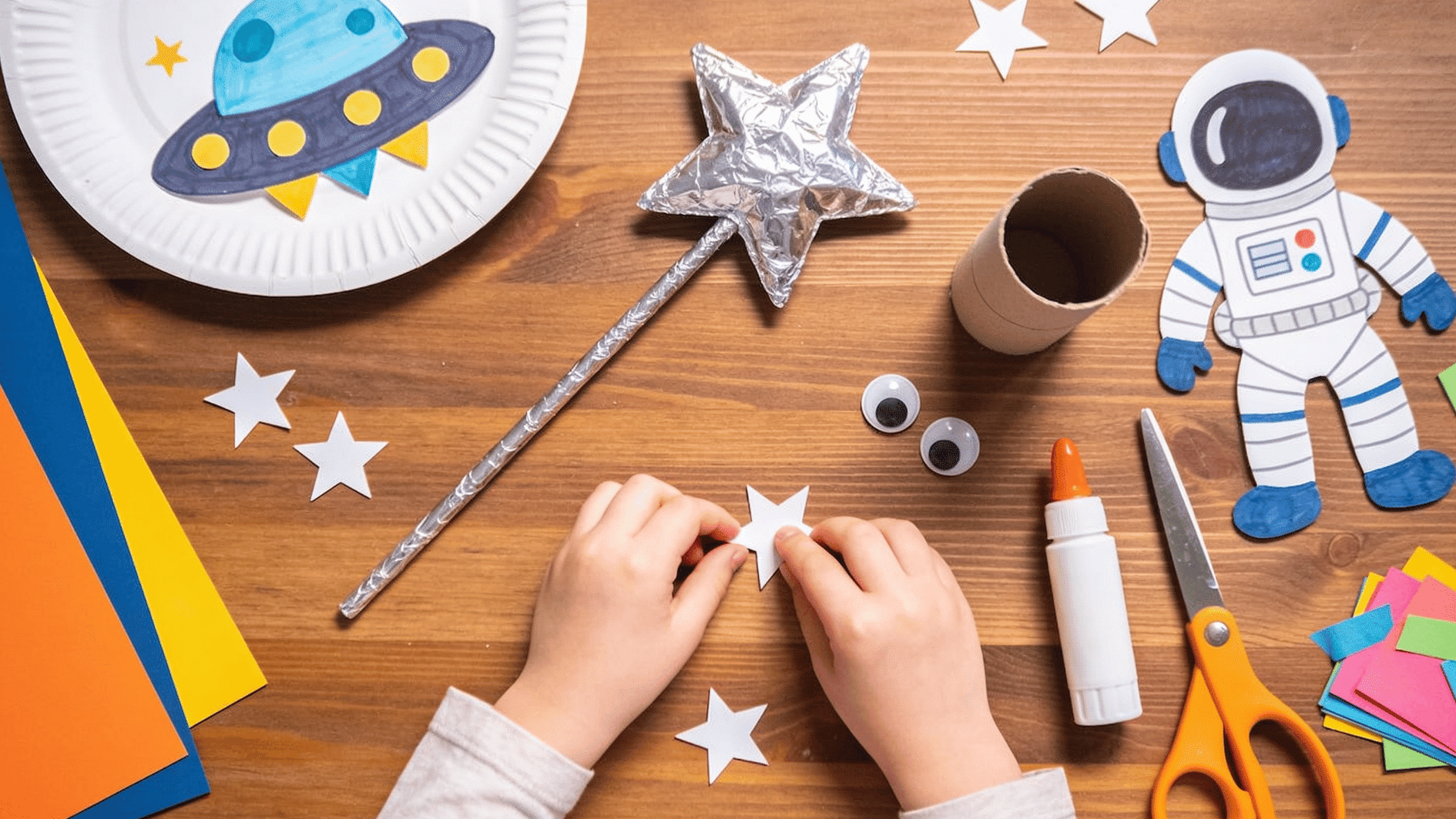 kids' space craft station on a wooden table — a half-finished paper plate UFO, a foil-wrapped star wand, a toilet paper roll astronaut, scattered star stickers, googly eyes, glue, scissors, and co