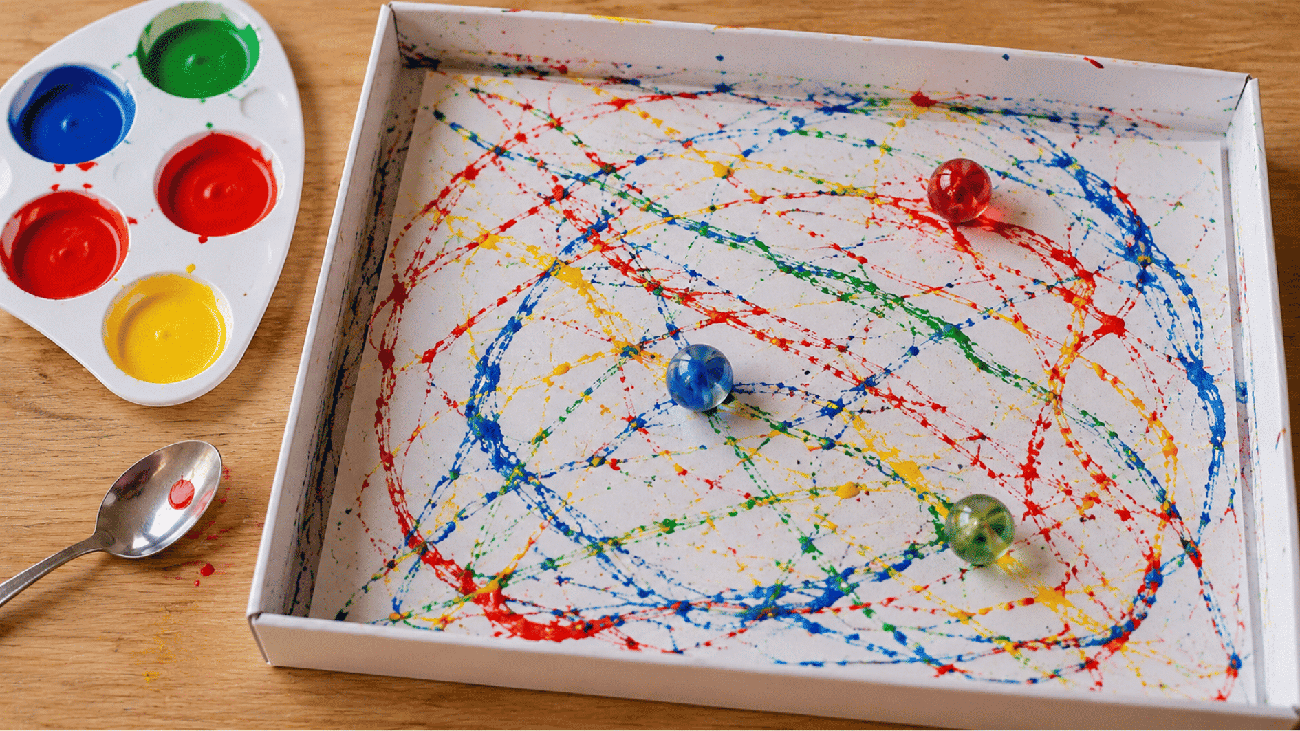 marble painting with red blue green and yellow trails inside a tray beside paint pots and spoon