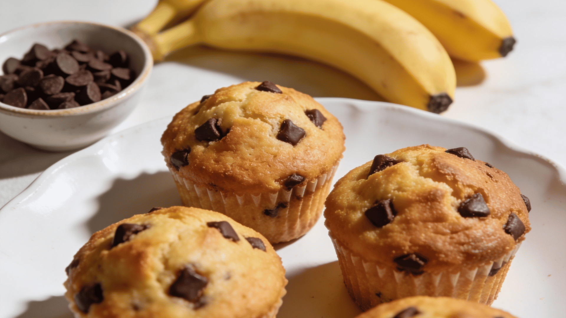 mini chocolate chip banana muffins on a white plate with mini chocolate chips and ripe bananas in the background