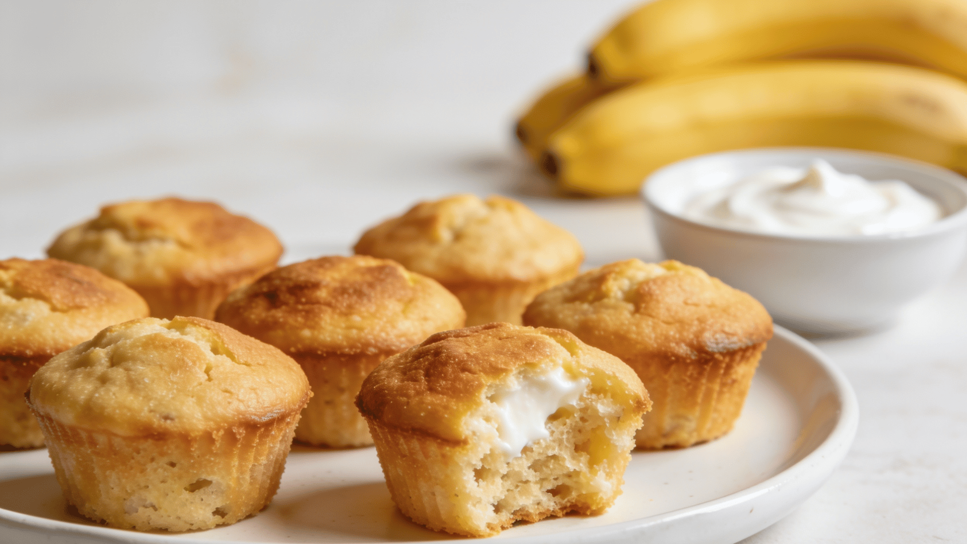 mini greek yogurt banana muffins on a white plate with plain yogurt and ripe bananas in the background