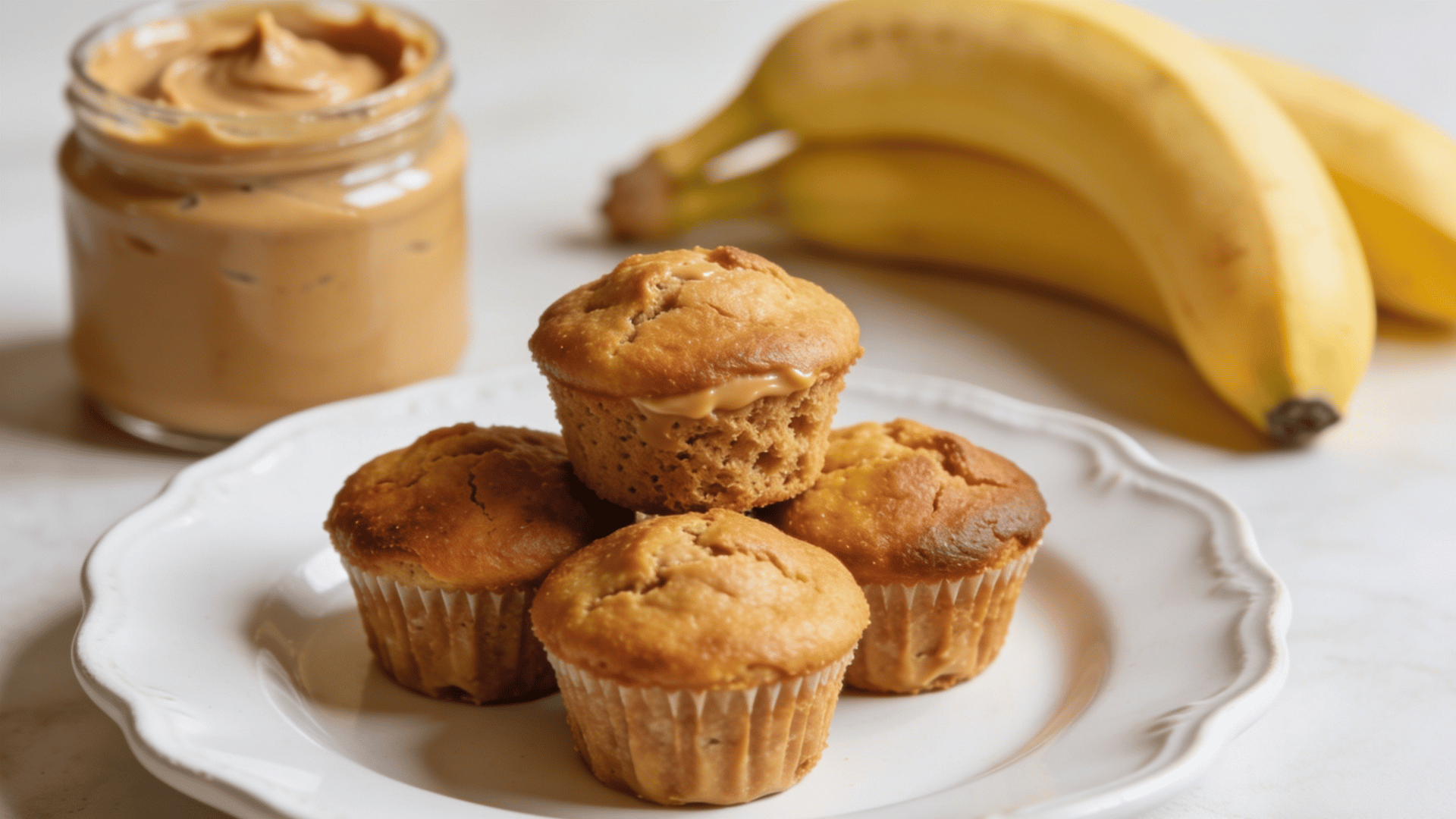 mini peanut butter banana muffins on a white plate with peanut butter and ripe bananas in the background