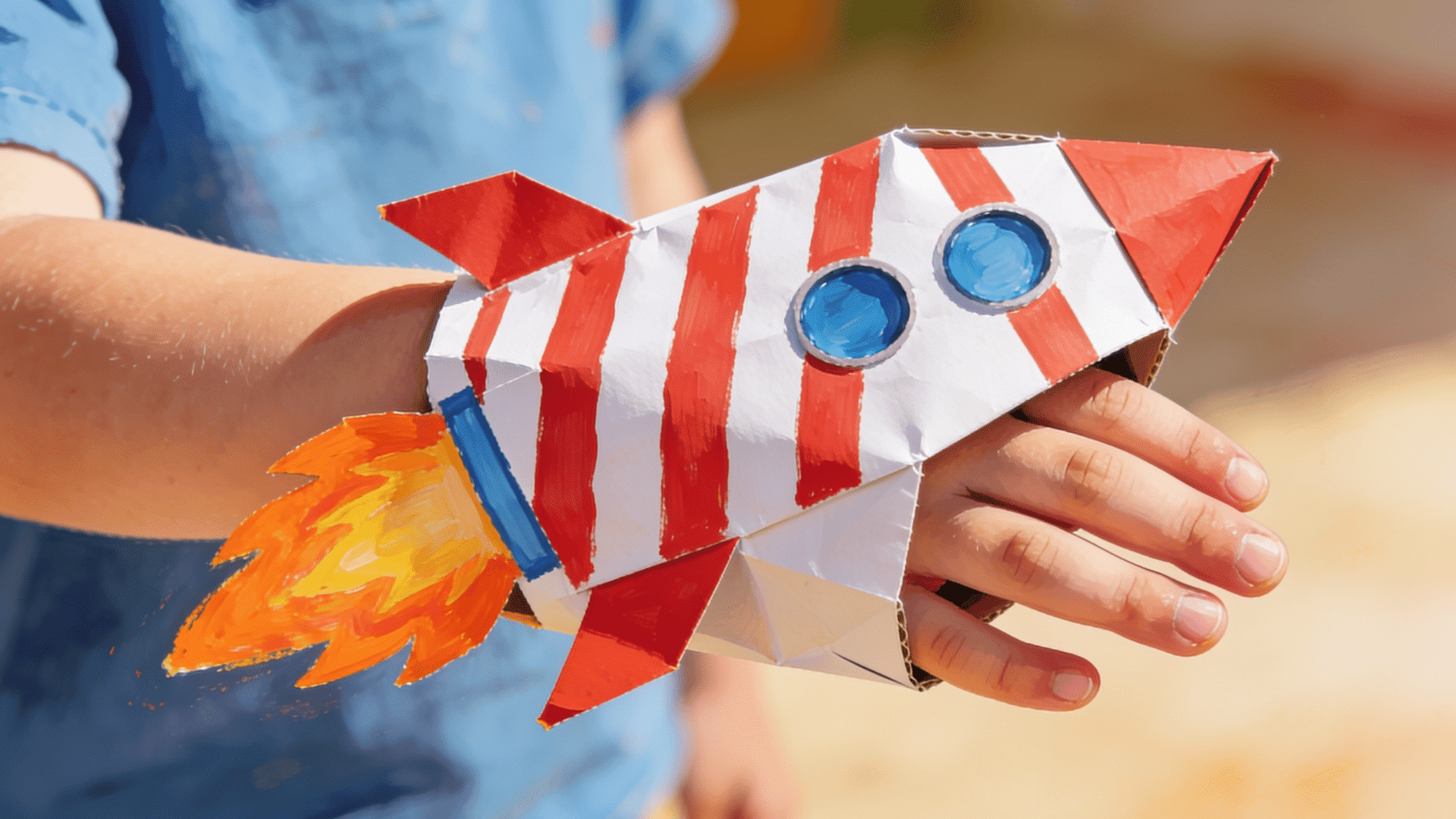 painted paper lunch bag puppet designed to look like a rocket with stripes, windows, and flame details, shown being worn on a child's hand
