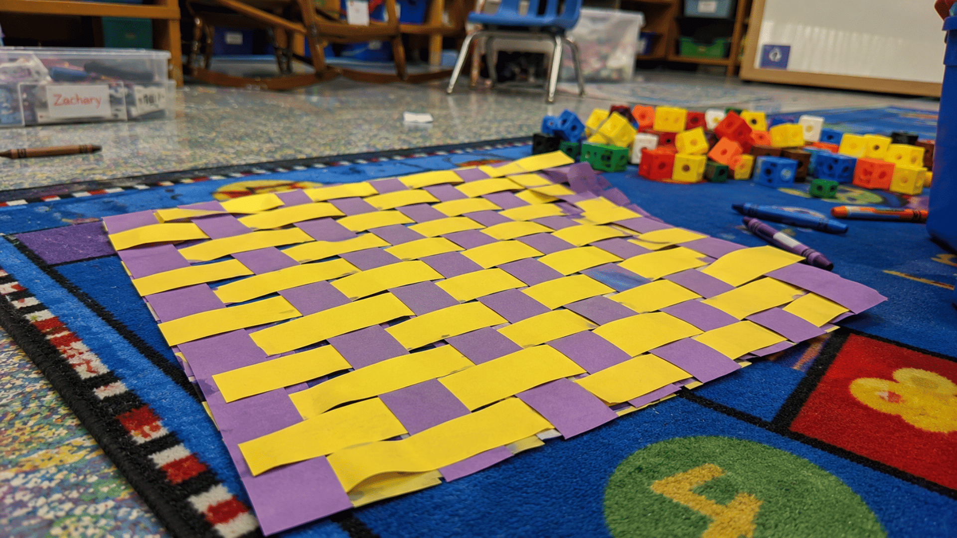 purple and yellow woven paper mat on a classroom rug surrounded by crayons and blocks