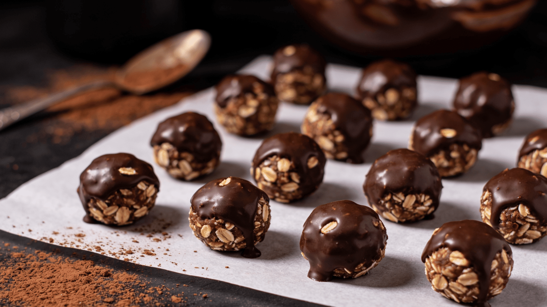 small chocolate oat energy bites arranged neatly on parchment paper, slightly glossy chocolate coating, oats visible on surface, cocoa powder dusting around, a spoon and mixing bowl in background