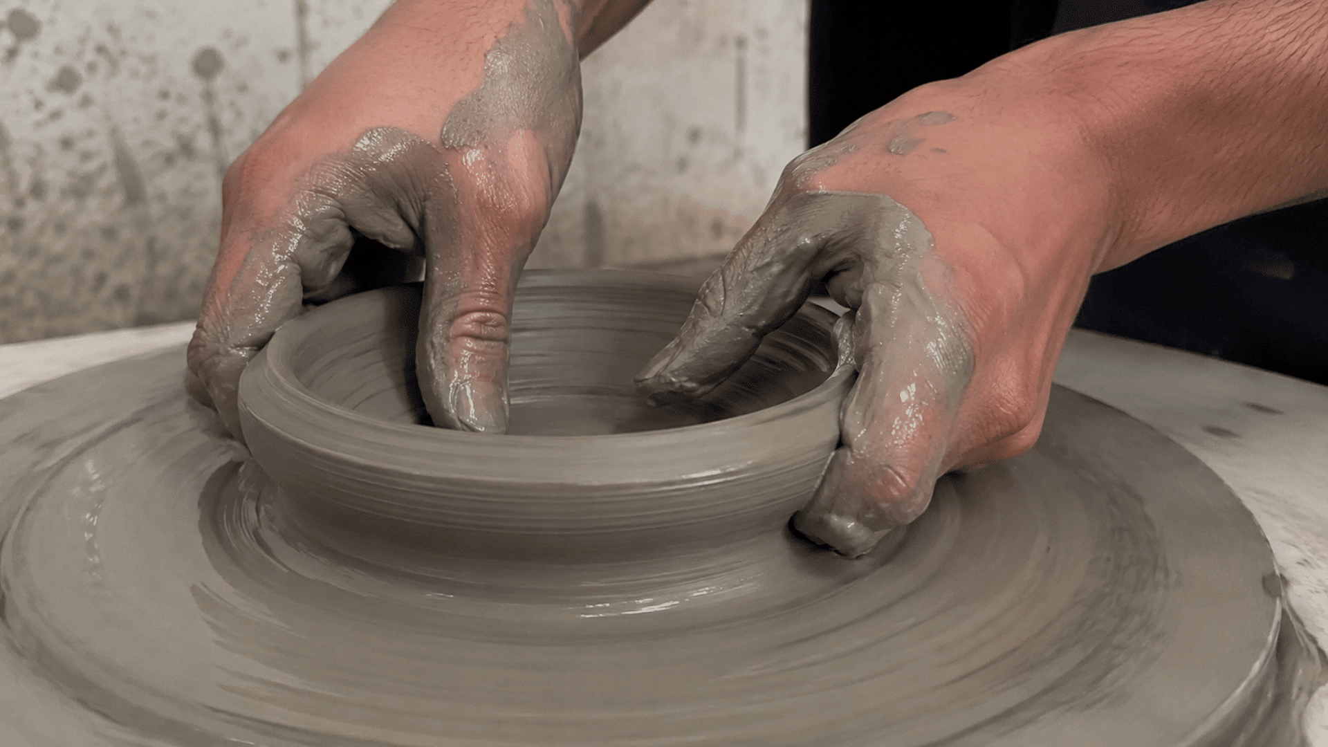Two clay-covered hands shaping a bowl from wet grey clay on a spinning pottery wheel during the plastic clay stage.