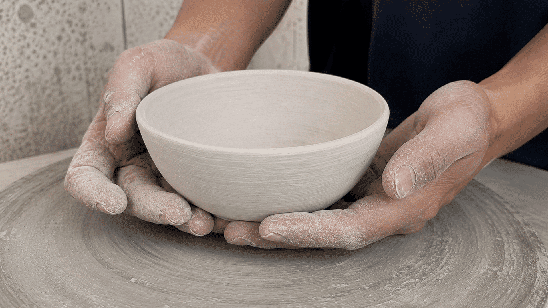 Two dusty hands gently cradling a pale, chalky bone-dry clay bowl on a pottery wheel before firing.