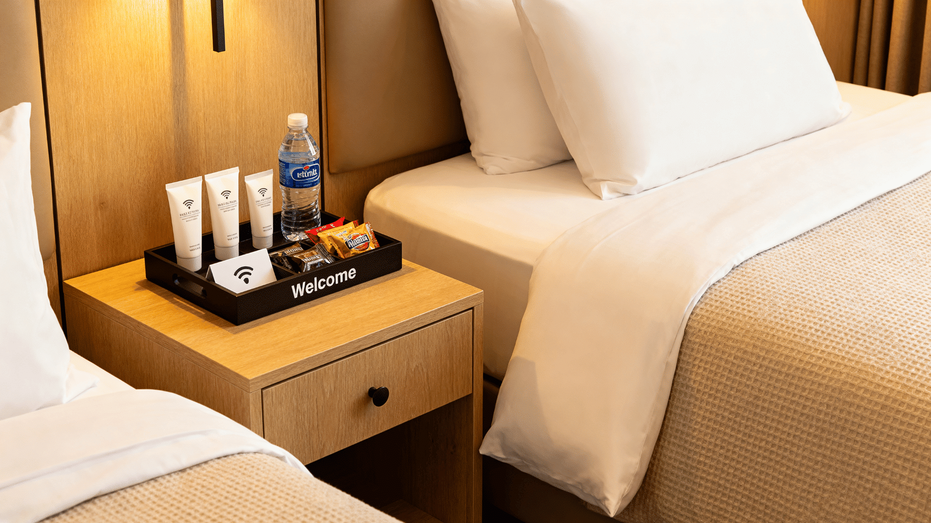 welcoming guest bedroom tray with travel toiletries bottled water snacks wifi note and neatly made bed behind it