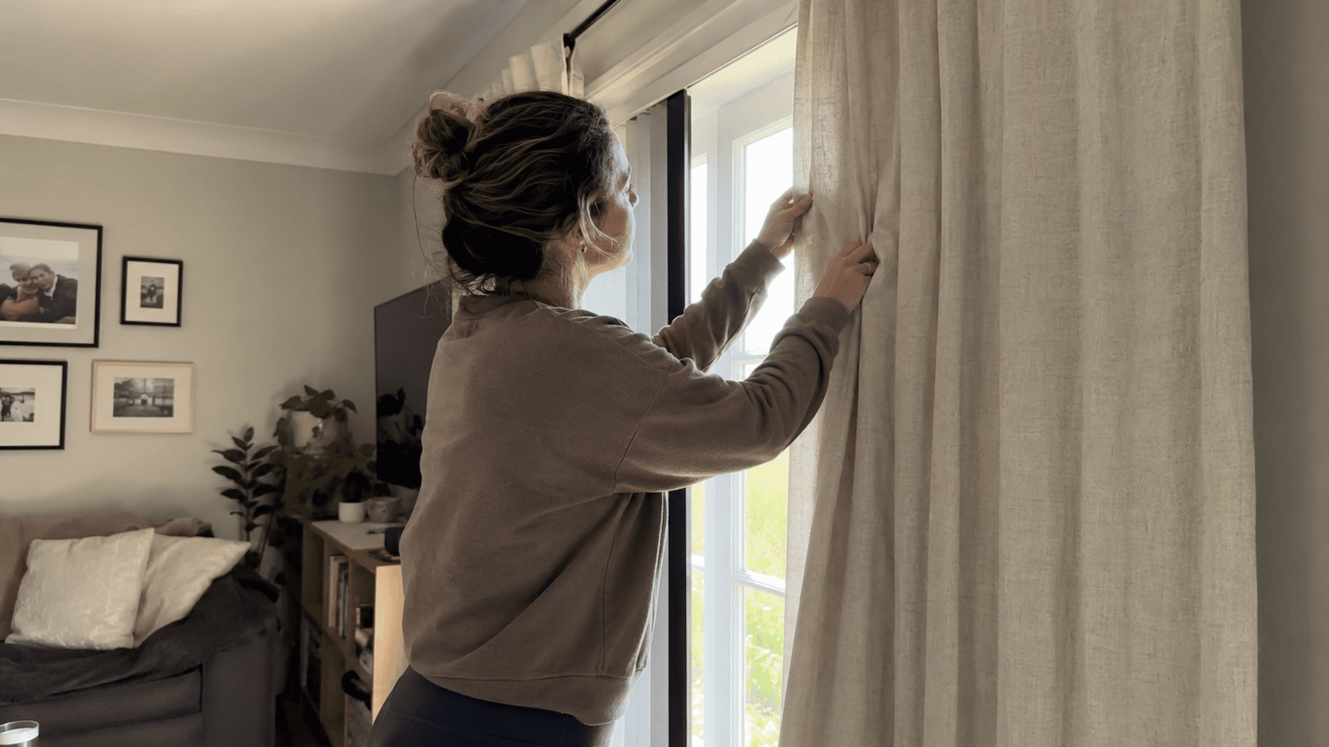 woman straightening linen curtains by a window with a cozy living room and gallery wall visible in the background