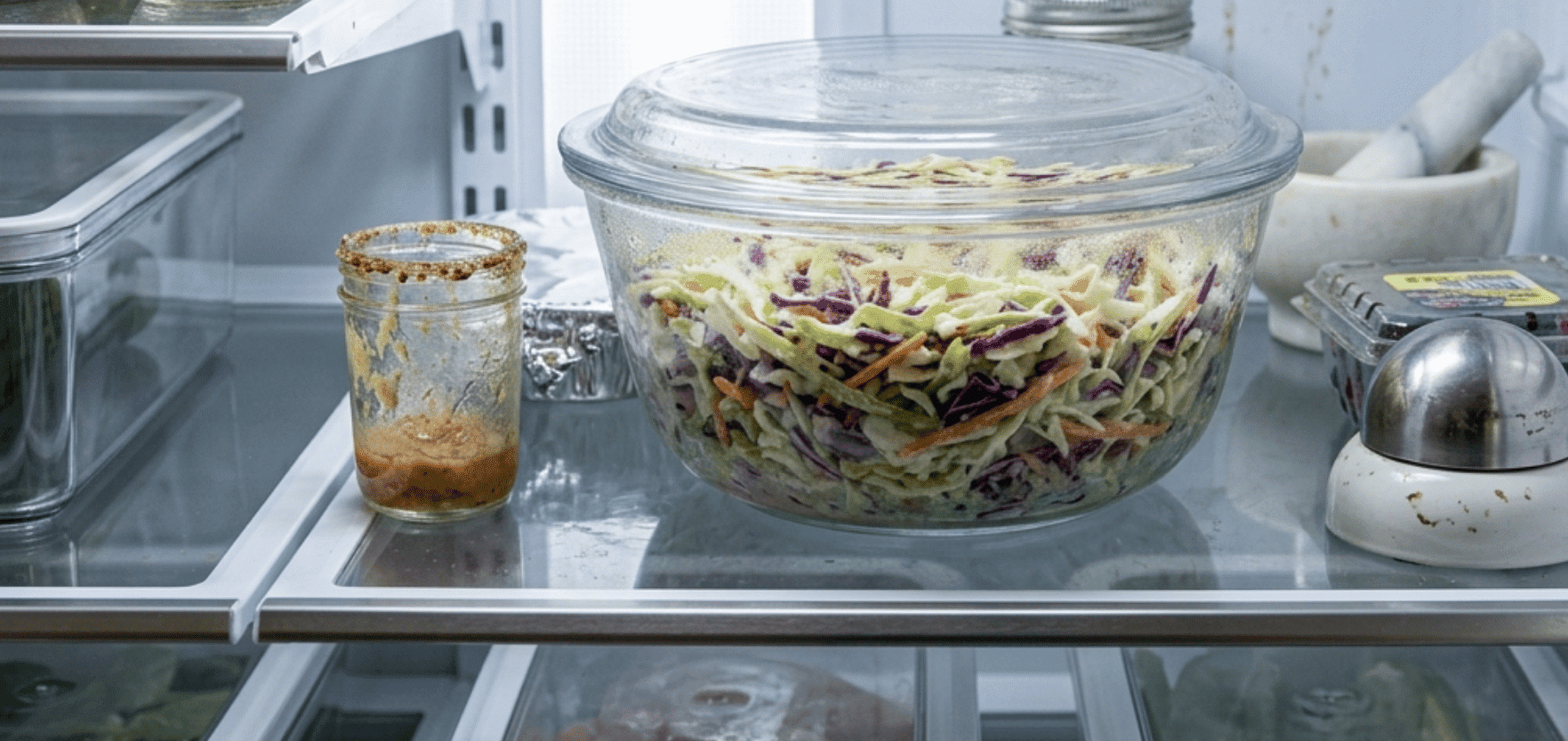 covered glass bowl of freshly dressed coleslaw resting inside a clean refrigerator before serving.png